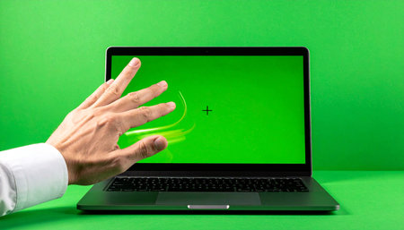 A person's hand gestures with a dynamic sweep over a laptop featuring a green screen, ready for custom content.の素材