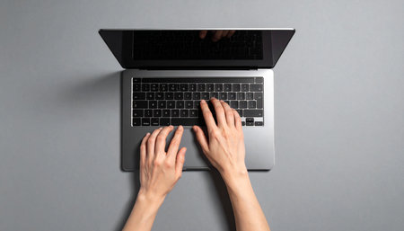 A focused individual types on a sleek laptop, captured from a top-down perspective against a minimalist gray background.の素材
