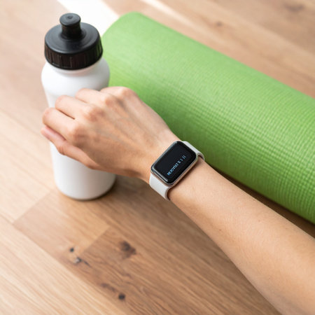 A moment of preparation before a yoga session. A woman checks her fitness goals on her smartwatch while reaching for her water bottle, with her yoga mat ready on the wooden floor.の素材