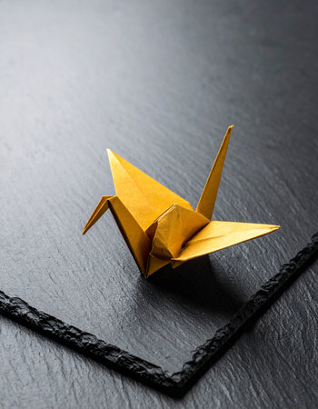 A single, elegant golden origami crane rests on a dark, textured slate surface.の素材