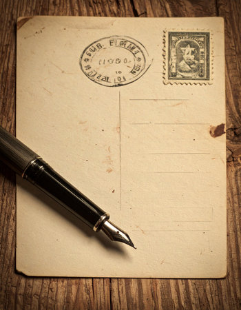 An antique postcard, yellowed with age and marked by a faint postmark, rests on a rustic wooden desk.の素材