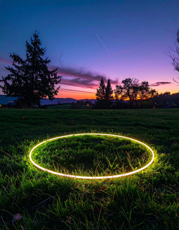 As twilight descends, a mysterious glowing ring of light illuminates a grassy field, creating a surreal and magical portal between the worlds of nature and technology.の素材
