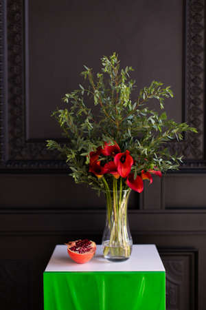 Bouquet of red calla lilies in a glass vase with a pomegranate and eucalyptus stands on the bedside table on a black backgroundの写真素材