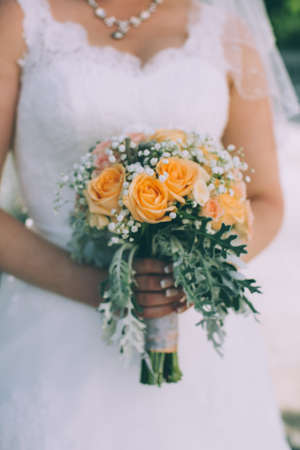 Beautiful bride with a wedding bouquet of orange roses against a background of green leaves in summer sunny dayの写真素材