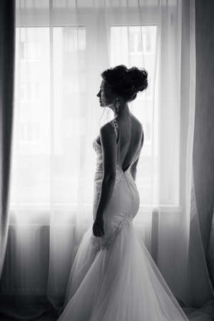 Silhouette of a beautiful bride in a traditional white wedding dress, stood by window.の写真素材