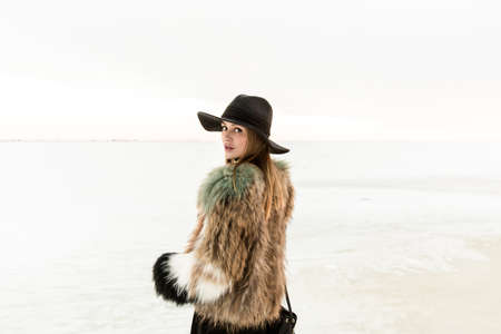 Portrait of a young beautiful blonde woman with hat, in a fur coat, outside, on the snow. A good dark day with beautiful shadows - a sunny winter day, sunset.の写真素材