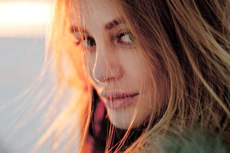 Portrait of a young beautiful blonde woman with hat, in a fur coat, outside, on the snow. A good dark day with beautiful shadows - a sunny winter day, sunset.の写真素材