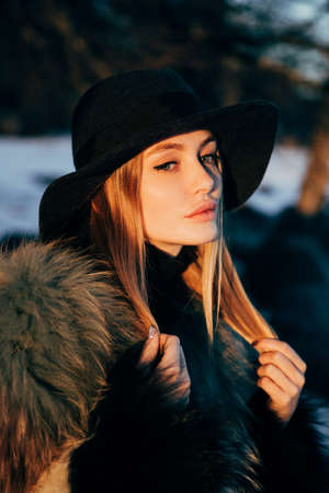 Portrait of a young beautiful blonde woman with hat, in a fur coat, outside, on the snow. A good dark day with beautiful shadows - a sunny winter day, sunset.の写真素材
