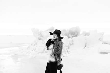 Portrait of a young beautiful blonde woman with hat, in a fur coat, outside, on the snow. A good dark day with beautiful shadows - a sunny winter day, sunset.の写真素材