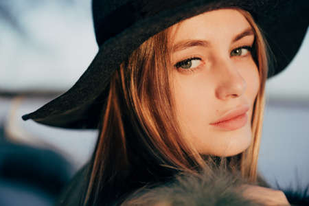 Portrait of a young beautiful blonde woman with hat, in a fur coat, outside, on the snow. A good dark day with beautiful shadows - a sunny winter day, sunset.の写真素材