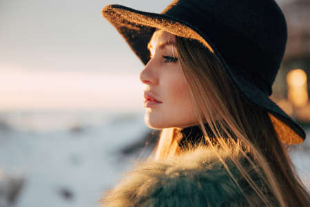Portrait of a young beautiful blonde woman with hat, in a fur coat, outside, on the snow. A good dark day with beautiful shadows - a sunny winter day, sunset.の写真素材