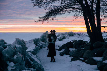 Elegant girl in fur coat and hat at sunset against frozen seaの写真素材