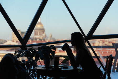 Silhouette of a woman in a cafe with a cup of coffee on a city backgroundの写真素材