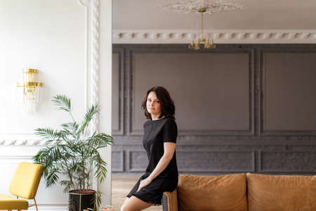 Woman in a black dress sits on the back of a sofa in the interiorの写真素材