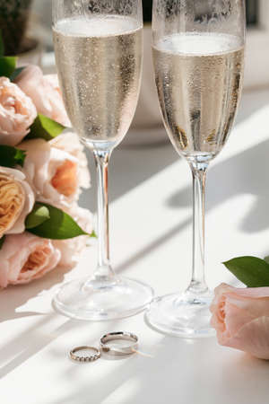 Wedding rings lie next to two glasses of champagne and a bouquet of roses in the sunの写真素材