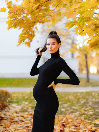 Young beautiful pregnant woman with dark hair in a black tight dress posing on an autumn meadow in the parkの写真素材