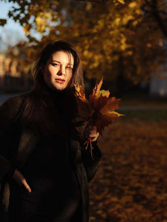 Young beautiful pregnant woman with dark hair in a black tight dress and coat posing on an autumn meadow in the parkの写真素材