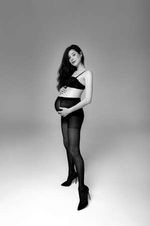 Pregnant brunette woman in pantyhose, bra and shoes stands on a gray backgroundの写真素材