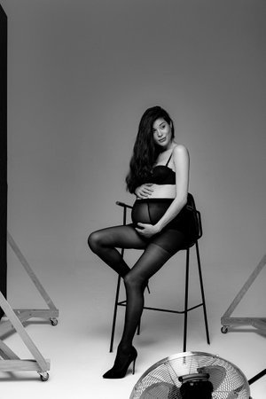 Pregnant brunette girl in pantyhose, bra and shoes on a high chair in a photo studioの写真素材