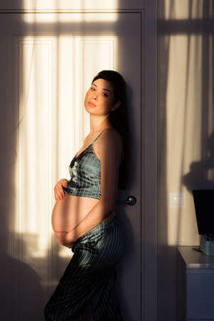 Pregnant woman in pajamas standing in the sun at homeの写真素材
