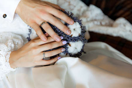 Bride and groom hold hands with rings on a bouquet of lavender with cottonの写真素材