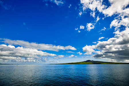 Volcanic island of Rangitoto in New Zealand seen across a expanse of blue waters and a background of a dazzling blue sky with a scattering of white cloudsの写真素材