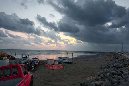 Fishing boats and a pickup on a beach at sunrise against a cloudy sky breaking into golden hues as the sun risesの写真素材