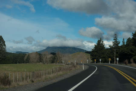 Tree lined road curving to the left under cloudy skies and a mountain in the backgroundの写真素材