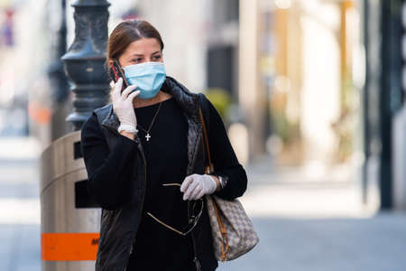 Vienna, Austria - April 04, 2020: Old woman wearing a mask on the street, protection against corona virus infection, using smartphone on a sunny day in springtimeのeditorial素材