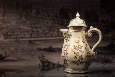 Closeup of a beautiful old coffee pot made of porcelain, painted with flowers standing on a reflective tableの写真素材