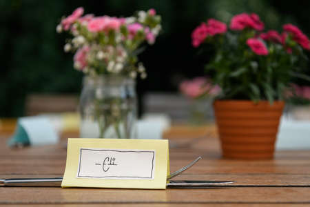 Hand written table name card and cutlery on a wooden table, flowers in the background, wedding, outdoorsの写真素材