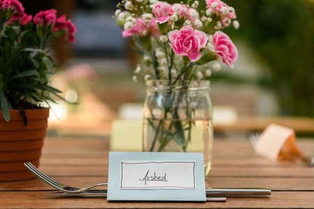 Hand written table name card and cutlery on a wooden table, flowers in the background, wedding, outdoorsの写真素材
