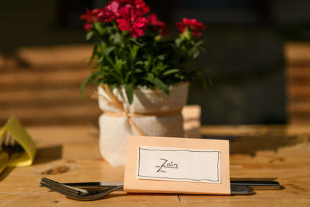 Hand written table name card and cutlery on a wooden table, flowers in the background, wedding, outdoorsの写真素材