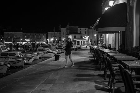 Cres, Croatia - October 24, 2022: The harbor of Cres in the night, empty chairs of a bar, woman on the phone, black and whiteのeditorial素材
