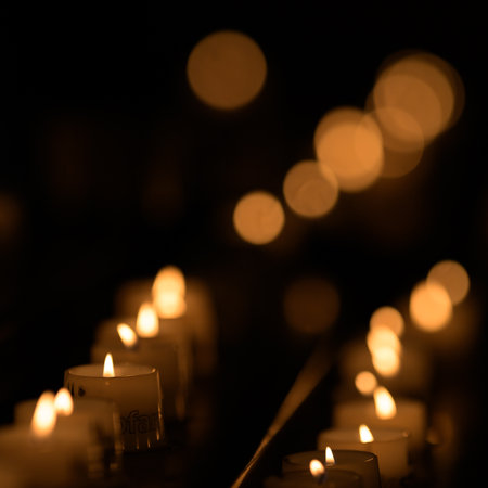 Burning votive candles with one flame in focus against dark background and beautiful bokeh of other flamesの写真素材