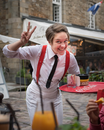 Saint-Malo, France - August 12, 2018: A waitress with white blouse red suspenders and black tie holds a tray making a victory sign.のeditorial素材