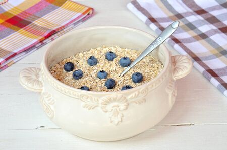 Decorative bowl of fine oat flakes with fresh fruit (blueberries, blackberries) on white wooden table with checkered dishtowelの写真素材