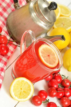 Fruit cherry tea with slice of lemon in glass mug, old teapot, red checkered tablecloth, fresh cherries and lemons in foregroundの写真素材