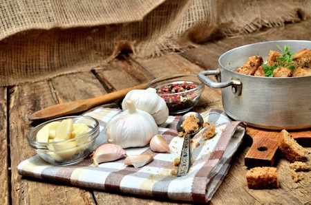 Close-up garlic soup in an old saucepan, fresh garlic bulbs, cloves, colored pepper, wooden spoon, croutons and a towel on a rustic wooden tableの写真素材