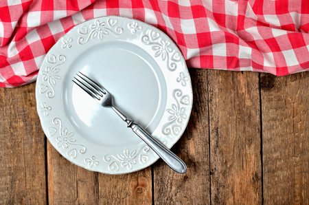 Fork in white decorative ceramic plate and red checkered tablecloth on top side on old vintage wooden table background - view from aboveの写真素材