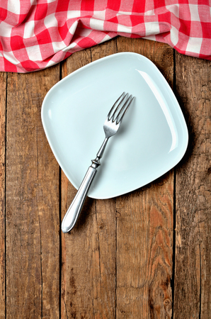 Decorative square plate, fork, and red checkered tablecloth on top side on old vintage wooden table background - view from above - vertical photoの写真素材