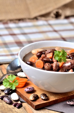 Bowl of bean soup with large beans on cutting board, carrots, parsley, marjoram, spoon and ladle, towel in background - vertical photoの写真素材