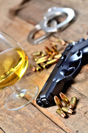 Glass of cognac, 9mm pistol, handcuffs and bullets on old wooden table - vertical photoの写真素材
