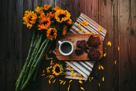 A bouquet of sunflowers, striped placemat, wooden board, cup of coffee, and brownies on a table.の写真素材