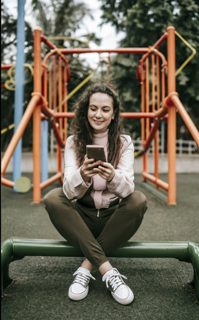 Woman sitting on the playground with a phone in her handsの素材