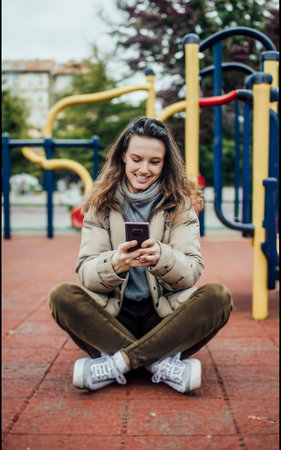 Woman sitting on the playground with a phone in her handsの素材
