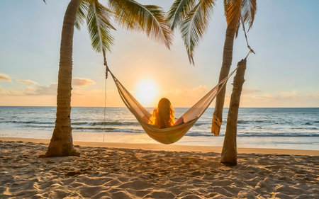 Back view of a young woman in a hammock on a tropical sandy beach during the sunsetの素材