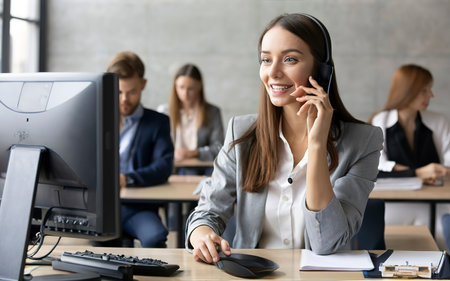 Young businesswoman talking on phone while sitting at desk in modern office. Smiling female customer service representative using headset for support.の素材
