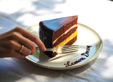 A person's hand with a silver ring uses a fork to cut into a rich chocolate cake slice. The cake has layers of chocolate sponge, cream, and bright yellow lemon filling, topped with glossy chocolate glaze.の写真素材