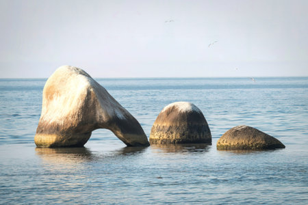 A unique rock formation with an archway stands in clear, shallow water under a bright sky. Three large stones are visible.の写真素材